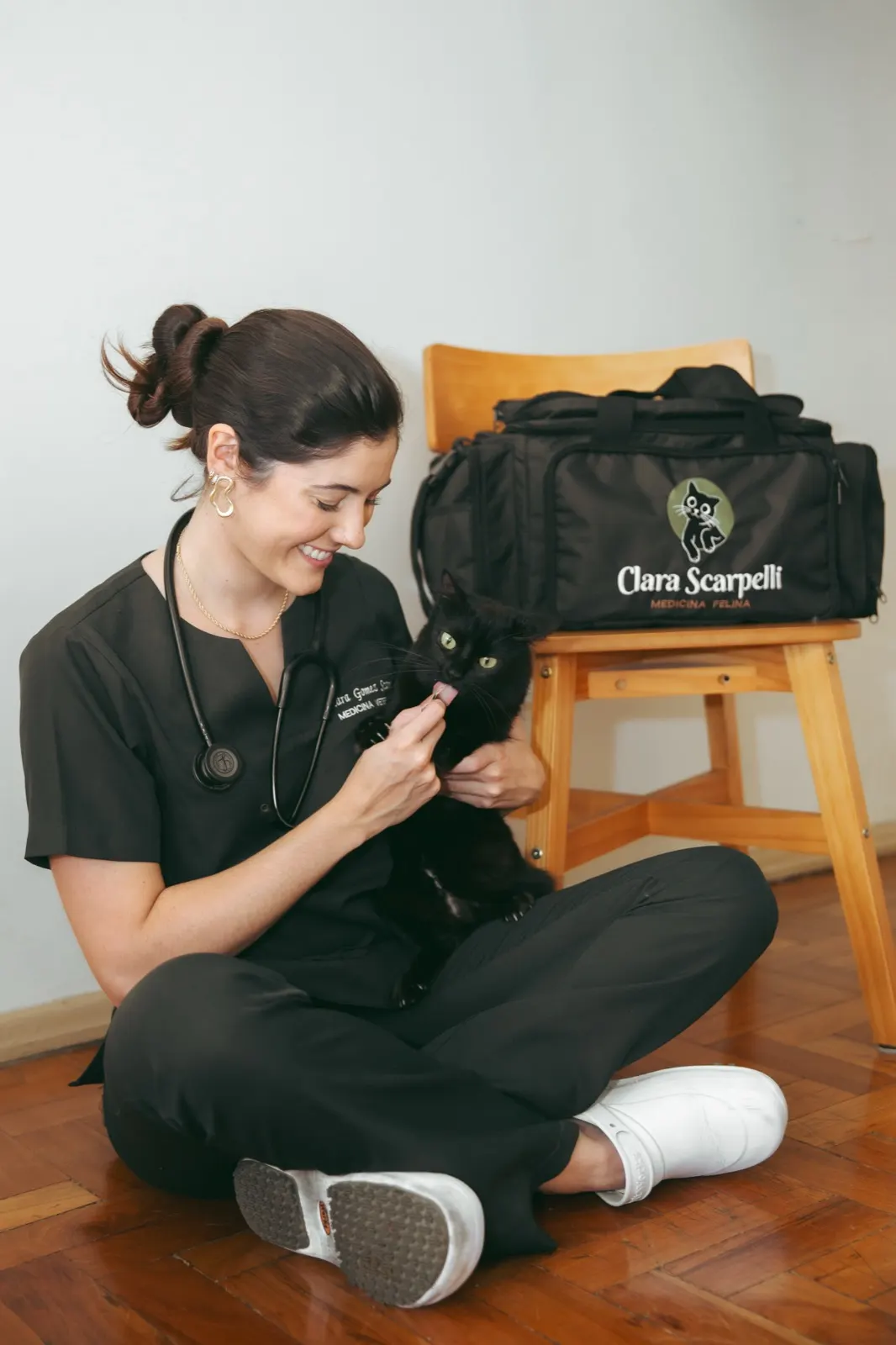 Dra. Clara Gomez Scarpelli sentada no chão oferecendo um petisco a um gato preto, ao lado da maleta da Clara Medicina Felina